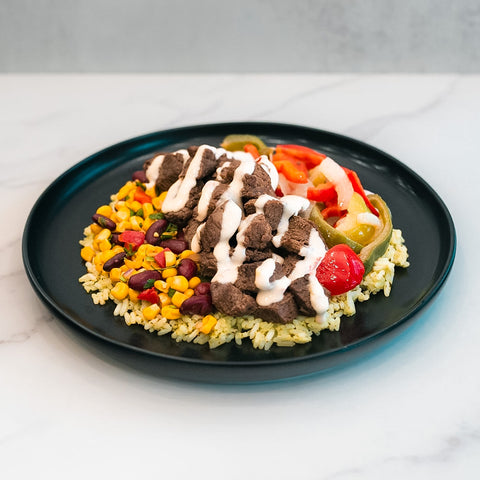Enjoy the Texas Queso Steak Bowl—a gluten-free, high-protein meal prep option featuring beef strips topped with white queso sauce, served over a bed of rice, corn, and kidney beans, and accompanied by sautéed bell peppers and onions.