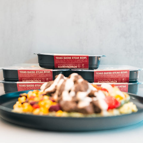 A plate of food featuring steak, corn, and beans is prominently displayed in the foreground, highlighting a delicious high-protein meal, with stacks of Texas Queso Steak Bowl packages visible in the background.
