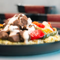 A close-up of a plate showcasing the Texas Queso Steak Bowl, topped with savory grilled beef strips, vibrant bell peppers, and a generous drizzle of creamy white sauce over a bed of rice. The high-protein dish is attractively presented with a softly blurred backdrop.