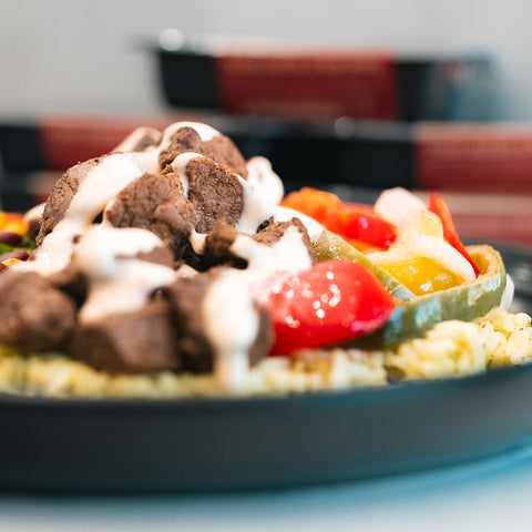 A close-up of a plate showcasing the Texas Queso Steak Bowl, topped with savory grilled beef strips, vibrant bell peppers, and a generous drizzle of creamy white sauce over a bed of rice. The high-protein dish is attractively presented with a softly blurred backdrop.