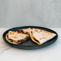 A black plate holds three Philly Cheesesteak Quesadillas, folded and filled with vegetables and cheese, displayed on a white marble surface against a light gray background—making them an ideal choice for a macro-friendly or high-protein meal prep.