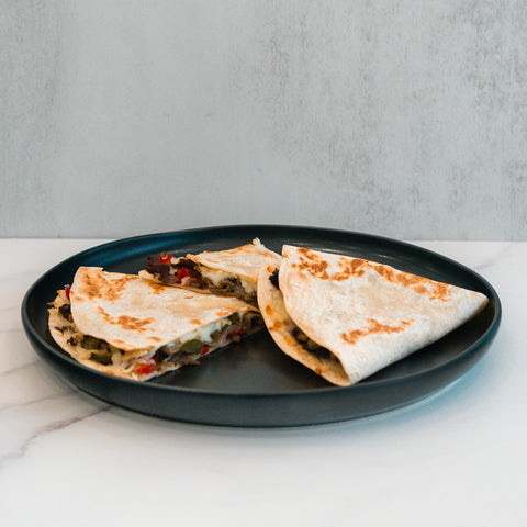 A black plate holds three Philly Cheesesteak Quesadillas, folded and filled with vegetables and cheese, displayed on a white marble surface against a light gray background—making them an ideal choice for a macro-friendly or high-protein meal prep.