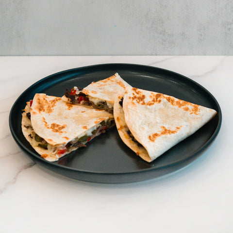 A black plate holds two Philly Cheesesteak Quesadillas—filled with vegetables and each cut in half—resting on a white marble surface with a light gray background, making them perfect for macro-friendly, high-protein meal prep.