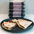 A black plate with two quesadilla halves is displayed in front of a stack of five meal containers labeled Philly Cheesesteak Quesadilla, showcasing this macro-friendly, high-protein meal prep as an ideal option.