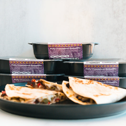Four Philly Cheesesteak Quesadilla meal prep packages are stacked in the background, while two quesadilla halves rest on a black plate in the foreground. The packaging displays purple labels with nutrition facts, showcasing Philly Cheesesteak Quesadilla as a high-protein, macro-friendly, and delicious meal option.
