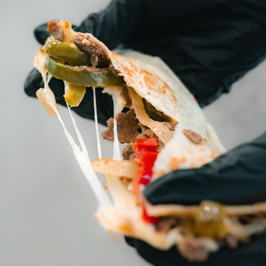 A gloved hand holds the Philly Cheesesteak Quesadilla, packed with tender beef, green peppers, onions, red peppers, and melted cheese stretching as it’s pulled apart—making it a delicious and macro-friendly option ideal for high-protein meal prep.