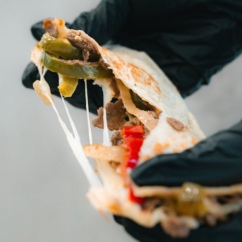 A gloved hand holds the Philly Cheesesteak Quesadilla, packed with tender beef, green peppers, onions, red peppers, and melted cheese stretching as it’s pulled apart—making it a delicious and macro-friendly option ideal for high-protein meal prep.