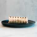 The "Golden Arches" Mac Daddy Wrap, a grilled burrito with char lines on a dark blue plate, delivers cheeseburger-inspired flavor for a macro-friendly lunch against a light, neutral background.
