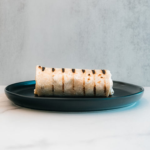 The "Golden Arches" Mac Daddy Wrap, a grilled burrito with char lines on a dark blue plate, delivers cheeseburger-inspired flavor for a macro-friendly lunch against a light, neutral background.