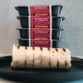 A grilled "The Golden Arches" Mac Daddy Wrap, a high-protein meal prep option, sits on a black plate with five black containers labeled with its name and offering cheeseburger-inspired flavor in the background.