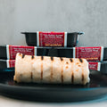 A "The Golden Arches" Mac Daddy Wrap rests on a black plate in front, with several packaged meal containers labeled the same stacked in the background.