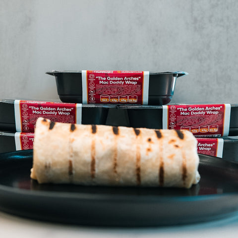 A "The Golden Arches" Mac Daddy Wrap rests on a black plate in front, with several packaged meal containers labeled the same stacked in the background.