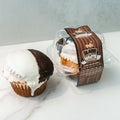 A Buffin Muffin - Black White Cookie sits beside a similar high-protein muffin in a clear plastic container with a brown and white label, displayed on a white surface.