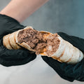 A person in black gloves holds "The Golden Arches" Mac Daddy Wrap, split to show its high-protein meal prep filling of ground meat and melted cheese for a cheeseburger-inspired taste.