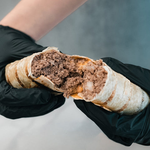A person in black gloves holds "The Golden Arches" Mac Daddy Wrap, split to show its high-protein meal prep filling of ground meat and melted cheese for a cheeseburger-inspired taste.