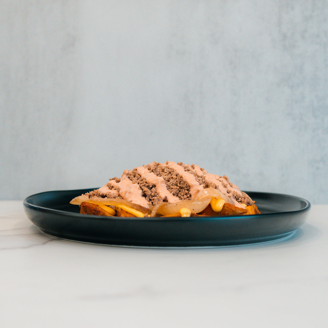 A black plate holds a slice of bread topped with a creamy spread, ground meat, and visible slices of pickles and cheese—reminiscent of the Animal Style West Coast Beef Bowl. This high-protein meal prep option stands out against the light gray background, set on a pristine white marble surface.