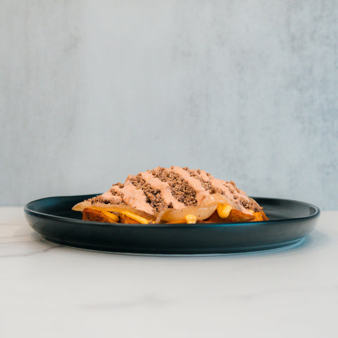 A black plate holds a slice of bread topped with a creamy spread, ground meat, and visible slices of pickles and cheese—reminiscent of the Animal Style West Coast Beef Bowl. This high-protein meal prep option stands out against the light gray background, set on a pristine white marble surface.