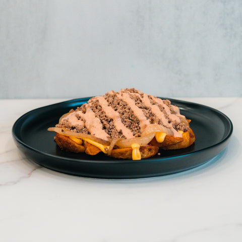 A serving of the Animal Style West Coast Beef Bowl features ground meat, melted cheese, diced onions, and a creamy sauce atop a waffle on a black plate—an ideal choice for anyone craving a macro-friendly take on this West Coast favorite.