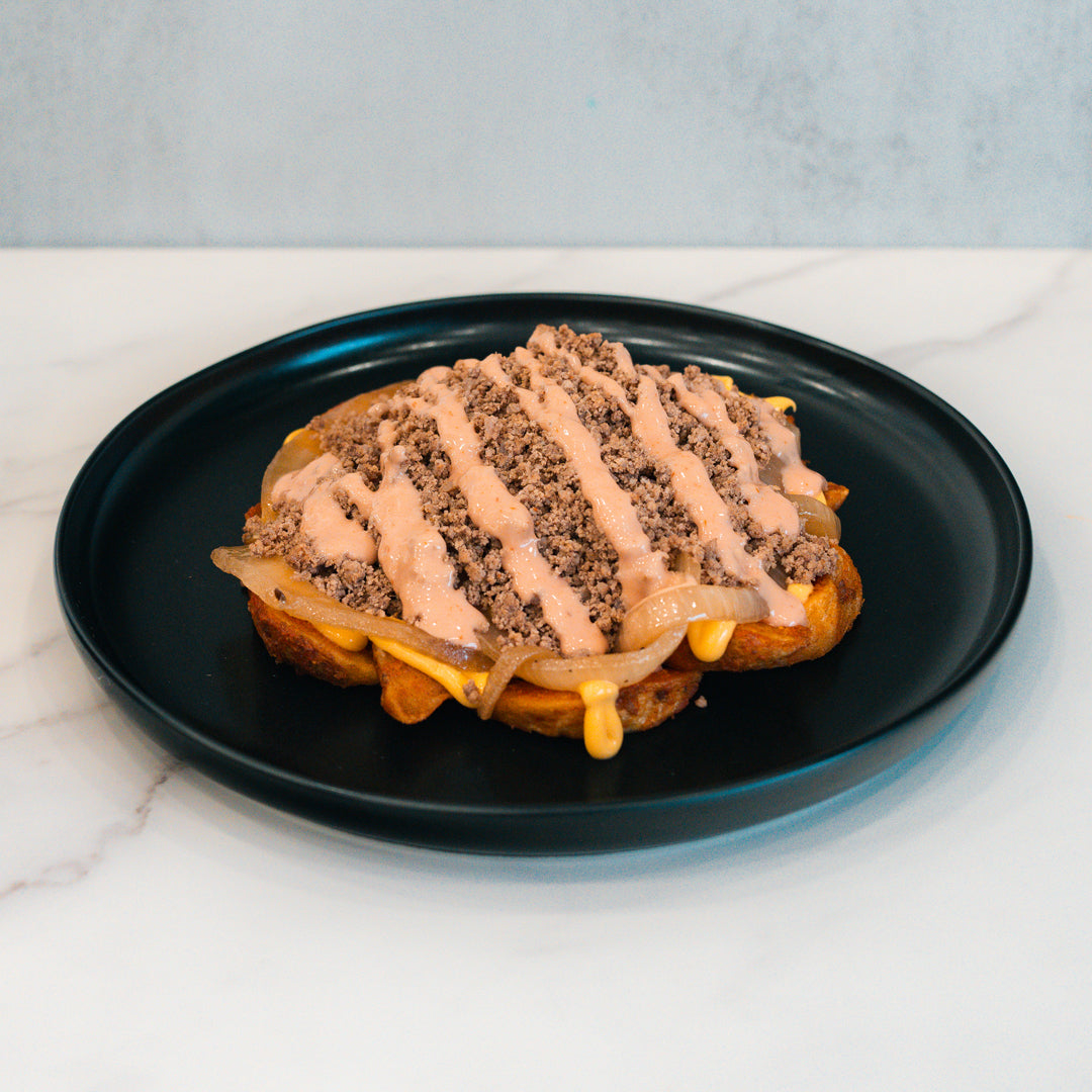 A macro-friendly Animal Style West Coast Beef Bowl: a waffle topped with sautéed onions, ground beef, melted cheese, and a drizzle of sauce, served on a black plate. Perfect for high-protein meal prep.