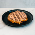 A macro-friendly Animal Style West Coast Beef Bowl: a waffle topped with sautéed onions, ground beef, melted cheese, and a drizzle of sauce, served on a black plate. Perfect for high-protein meal prep.