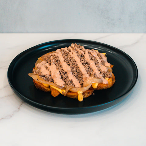 A macro-friendly Animal Style West Coast Beef Bowl: a waffle topped with sautéed onions, ground beef, melted cheese, and a drizzle of sauce, served on a black plate. Perfect for high-protein meal prep.