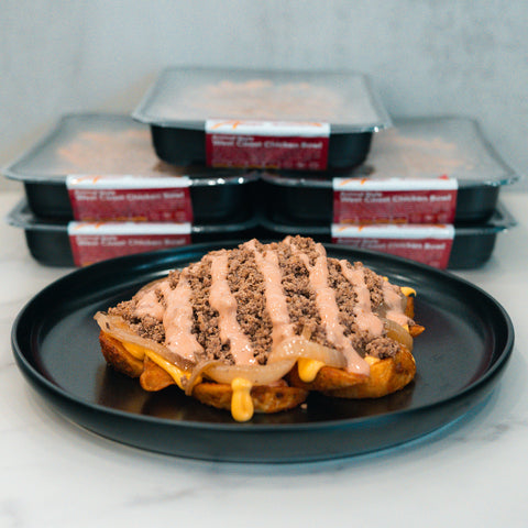 A black plate holds a waffle topped with ground meat, cheese, and sauce for a delicious macro-friendly twist. In the background, four packaged ready-made Animal Style West Coast Beef Bowl meals are stacked, perfect for high-protein meal prep.