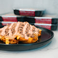 A close-up of the Animal Style West Coast Beef Bowl topped with crumbled chocolate, drizzled with two sauces, and served on a black plate. Blurry meal prep containers are stacked in the background, hinting at high-protein convenience.