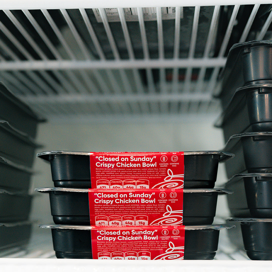 Three black Closed on Sunday Crispy Chicken Bowl containers with red labels are stacked in a white freezer, showcasing a macro-friendly lunch. Additional containers are visible to the right.