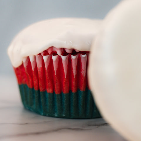 A close-up of the Red, White Blue Buffin Muffin—high in protein, with thick white icing and a red and dark green striped wrapper. Another similarly frosted muffin appears partially in the blurred background.
