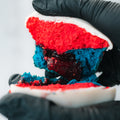 A close-up of hands in black gloves holding a Red, White Blue Buffin Muffin, featuring red and blue cake layers, white icing, and a dark berry filling in the center.