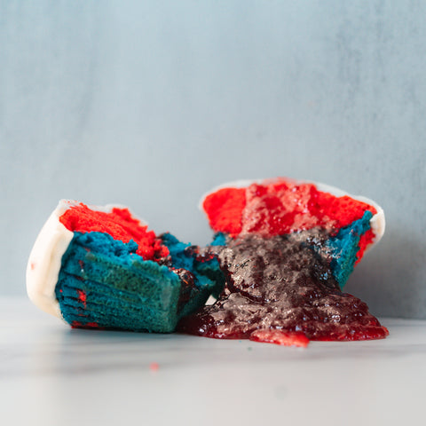 A broken Red, White Blue Buffin Muffin with red and blue sponge and white frosting is split open, showing a dark, gooey filling that has spilled out. The background is plain light gray.