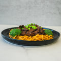 A black plate of Lo Mein Teriyaki Steak Subscription features high-protein noodles, sliced teriyaki steak, chopped green onions, and two steamed broccoli florets served on a light surface.