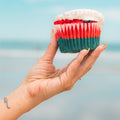 A hand with a small tattoo holds a Red, White Blue Buffin Muffin, featuring patriotic red, white, and blue layers plus white icing, against a blurred beach background.