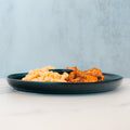 A dark plate on a white surface features Buffalo Mac N Chicken—macro-friendly comfort food with macaroni and cheese beside seasoned baked chicken—set against a light blue background.
