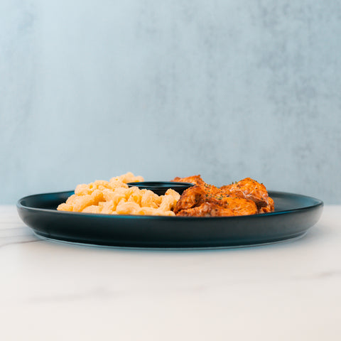 A dark plate on a white surface features Buffalo Mac N Chicken—macro-friendly comfort food with macaroni and cheese beside seasoned baked chicken—set against a light blue background.