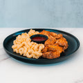 Buffalo Mac N Chicken features seasoned grilled chicken over mac and cheese, paired with a red dipping sauce on a black plate. This high-protein meal prep is ideal for those who love macro-friendly comfort food.