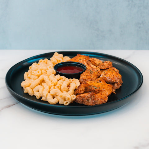 Buffalo Mac N Chicken features seasoned grilled chicken over mac and cheese, paired with a red dipping sauce on a black plate. This high-protein meal prep is ideal for those who love macro-friendly comfort food.