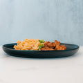 A black plate with spiral pasta, grilled chicken, and green herbs showcases the BBQ Chicken Mac Bowl—an inviting healthy meal prep option from Hummus Fit—set on a light surface with a blue-gray background.