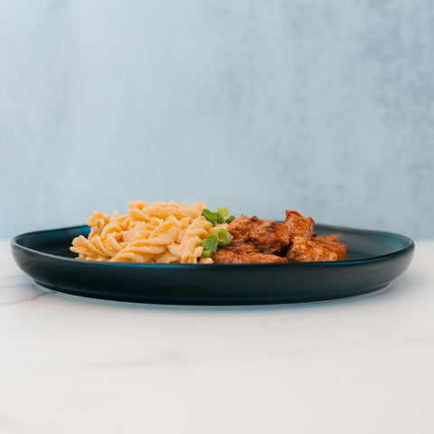 A black plate with spiral pasta, grilled chicken, and green herbs showcases the BBQ Chicken Mac Bowl—an inviting healthy meal prep option from Hummus Fit—set on a light surface with a blue-gray background.
