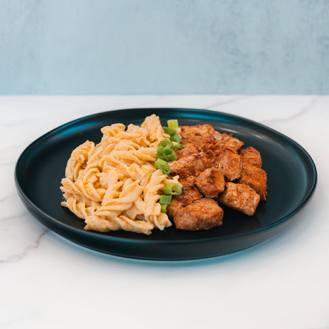 A black plate holds the BBQ Chicken Mac Bowl, a healthy meal prep featuring pasta with creamy sauce, seasoned chicken, and green onions. The meal sits on a white marble surface against a light blue background.