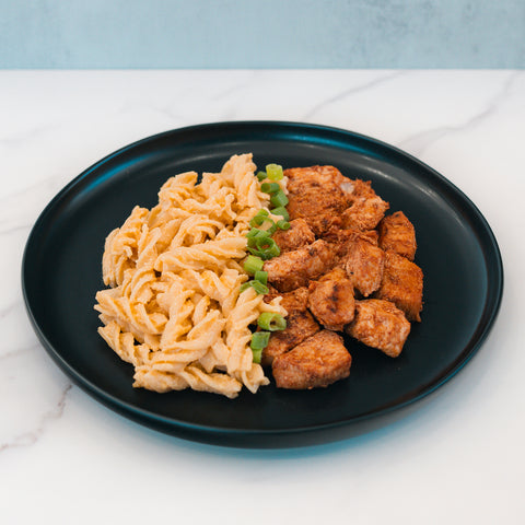 A black plate with BBQ Chicken Mac Bowl—creamy pasta topped with green onions—on a white marble surface, offers a nutritious meal prep option from Hummus Fit.