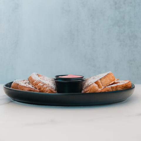 Three slices of Strawberry Protein French Toast, dusted with powdered sugar, are arranged on a black plate with a small cup of syrup in the center. The setting is minimal, with a light background and a marble surface—perfect for those seeking a low sugar breakfast without sacrificing indulgence.