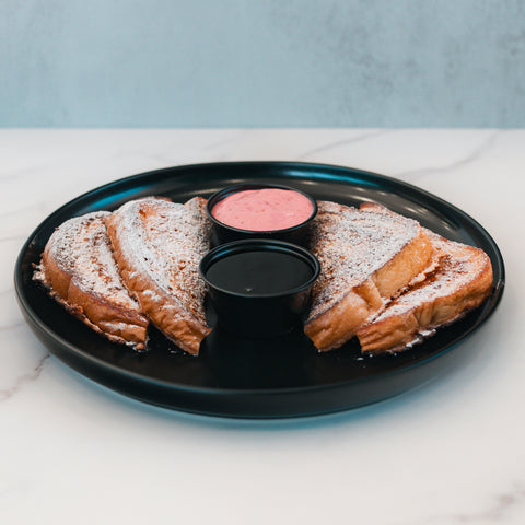 Four slices of Strawberry Protein French Toast, dusted with powdered sugar, are arranged on a black plate and served with two small cups of dipping sauces—one pink and one dark—on a white marble surface. This delicious twist on the classic is the perfect low sugar breakfast option.