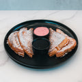 Three slices of Strawberry Protein French Toast, dusted with powdered sugar, are arranged on a black plate with two small containers of dipping sauce—one pink and one dark—on a white marble surface. Perfect for a low sugar breakfast, this delicious dish offers both flavor and nutrition.