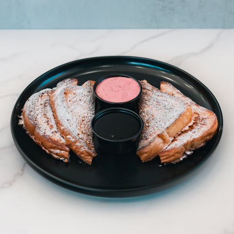 Three slices of Strawberry Protein French Toast, dusted with powdered sugar, are arranged on a black plate with two small containers of dipping sauce—one pink and one dark—on a white marble surface. Perfect for a low sugar breakfast, this delicious dish offers both flavor and nutrition.