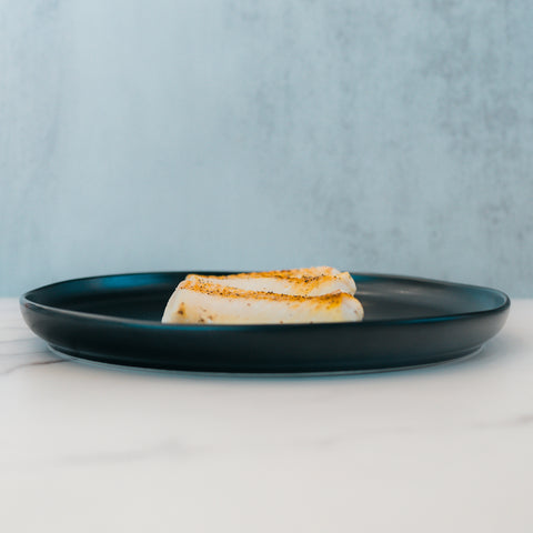 Two pieces of grilled halloumi cheese on a dark plate rest on a light marble surface with a pale blue background—ideal for meal prep or pairing with Competition Approved Oven Baked Cod 1lb for a protein-rich, balanced meal.
