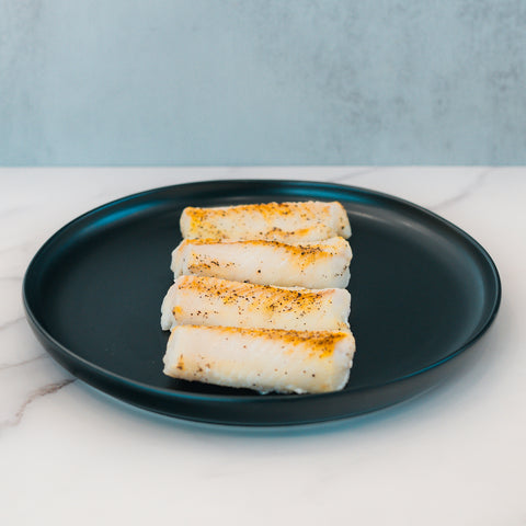 Three pieces of Competition Approved Oven Baked Cod 1lb are neatly arranged in a row on a black plate atop white marble—a delicious, high-protein option for meal prep.