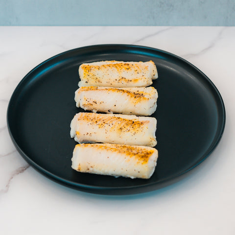 Four pieces of Competition Approved Oven Baked Cod 1lb arranged on a round black plate atop white marble—ideal for high-protein meal prep.
