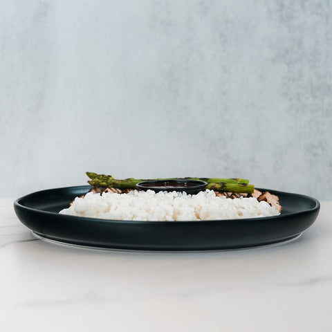 The Arnold 2022 Bowl features white rice, asparagus, grilled chicken, and a side of sauce in a black plate—perfect for a healthy lunch, presented on a white surface with a light gray background.