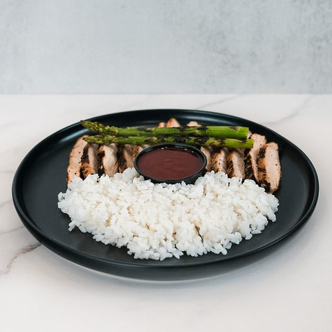 The Arnold 2022 Bowl features white rice, sliced grilled chicken breast, grilled asparagus, and a dark dipping sauce—ideal for healthy lunches or chicken and rice meal prep—all beautifully presented on a sleek black plate.